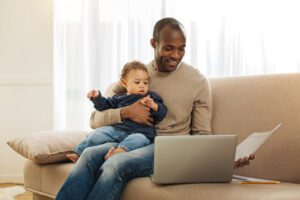 homem com bebê no colo segurando uma folha e um computador no sofá.