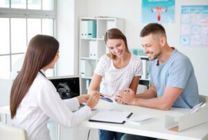 Casal hétero branco em consulta médica com uma doutora branca. Estão com expressões de felicidade.